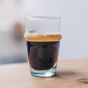 Può includere: Un bicchiere di vetro trasparente riempito di caffè nero sormontato da uno spesso strato di crema marrone chiaro. Il bicchiere presenta un distinto rilievo orizzontale vicino al bordo superiore e della condensa sulla superficie interna.