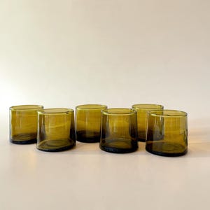 May include: A set of six short, cylindrical drinking glasses made of translucent olive green glass. Each glass features a smooth, slightly tapered shape with a thick, solid base and a simple, unadorned rim, arranged in a cluster.