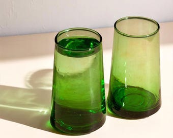 Peut inclure: Deux verres à boire coniques en verre vert translucide. Les verres présentent une forme cylindrique lisse et légèrement évasée avec une base épaisse, mettant en valeur une teinte émeraude vibrante qui capte la lumière pour créer des ombres.