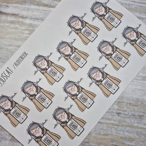 Könnte beinhalten: Bogen mit Aufklebern, die eine Zeichentrickfigur zeigen, die ein Hörbuch hört. Die Figur trägt Kopfhörer und hält ein Tablet. Das Tablet zeigt ein Podcast-Bild. Die Figur ist in eine beige Decke gehüllt. Der Text "MUSCAT / AUDIOBOOK" ist aufgedruckt.