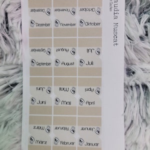 Könnte beinhalten: Ein Blatt mit 12 beigefarbenen Aufklebern mit einem Totenkopf-Motiv und den Monaten des Jahres in deutscher Sprache. Die Aufkleber sind für die Verwendung in einem Planer oder Tagebuch gedacht.