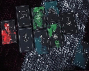 May include: A spread of tarot cards with black backgrounds and white illustrations. The cards are arranged in a circle with the top card being the "Three of Cups". The cards are on a dark gray surface.