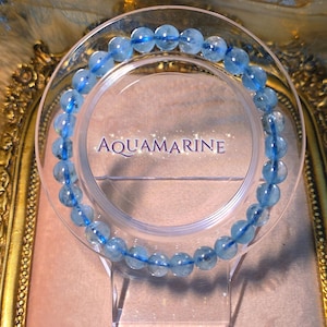 May include: A bracelet made of light blue aquamarine beads. The beads are round and strung together on a clear elastic cord. The bracelet is displayed on a clear acrylic stand with the word "Aquamarine" printed on it.