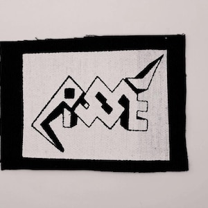 Può includere: Una toppa rettangolare in tessuto con bordo nero e centro bianco. Il centro mostra un design grafico nero stilizzato che ricorda la parola "RIOT" in un carattere angolare, appuntito e a blocchi con un distinto effetto tridimensionale.