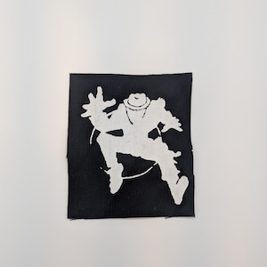 May include: A square black fabric patch featuring a white screen-printed graphic of a figure wearing a hat, depicted in a dynamic, jumping pose with one arm extended upward and legs bent, showcasing a high-contrast silhouette design.