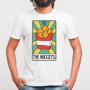 May include: White t-shirt with a graphic of a tarot card. The card is titled "The Nuggets" and features a red and white box of chicken nuggets. The card number is XII.