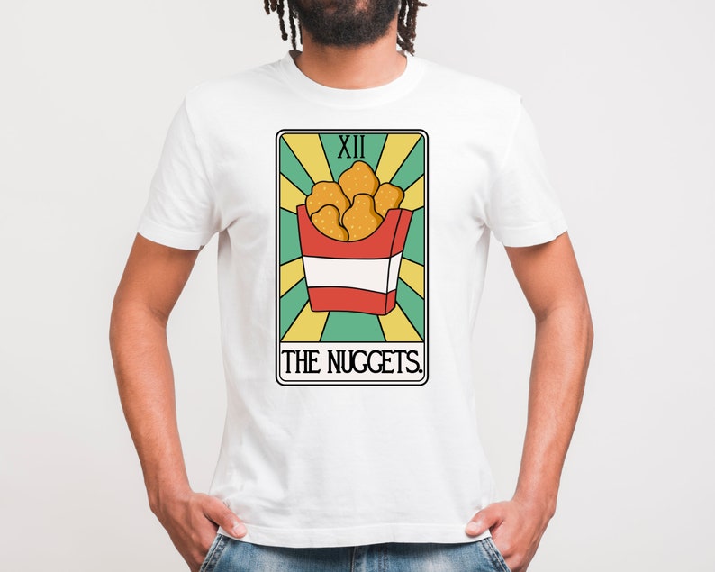 May include: White t-shirt with a graphic of a tarot card. The card is titled "The Nuggets" and features a red and white box of chicken nuggets. The card number is XII.