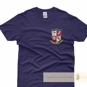 Könnte beinhalten: Ein marineblaues T-Shirt mit einem Wappen, das einen Totenkopf, einen Regenschirm, einen Blitz und eine Schere zeigt. Das Wappen ist von einem goldenen Band mit dem Text "Ut Palam Firma." umgeben.