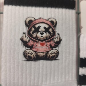 Könnte beinhalten: Weiße Socken mit schwarzen Streifen am oberen Rand. Die Socken zeigen eine Grafik eines Teddybären mit rosa Kapuzenpullover, durchkreuzten Augen und Mittelfinger. Der Text "I ❤️ Valentinstag" ist aufgedruckt.