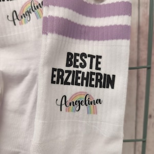 May include: White socks with lavender stripes and the text "BESTE ERZIEHERIN" and "Angelina" with a rainbow graphic. The socks are held together with a wooden clip.