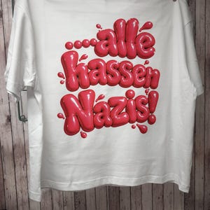 Könnte beinhalten: Ein weißes T-Shirt mit kurzen Ärmeln und einem großen, glänzenden, dreidimensionalen roten Grafikdruck auf der Vorderseite. Das Design zeigt den Text "alle hassen Nazis!" in einer abgerundeten, ballonartigen Schriftart mit kleinen roten Farbtropfen.