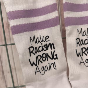 Könnte beinhalten: Weiße Socken mit lavendelfarbenen Streifen am oberen Rand. Die Socken tragen den Schriftzug "Make Racism WRONG Again!" in schwarzer Schrift. Die Socken hängen an einem Metallgitter mit einer Holzklammer.