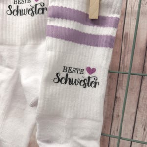Könnte beinhalten: Ein Paar weiße Socken mit lila Streifen und dem Text "Beste Schwester" mit einem Herzsymbol.