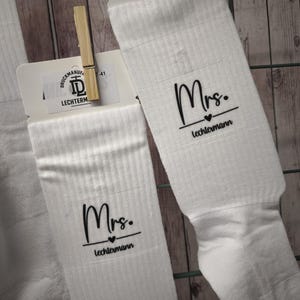 Könnte beinhalten: Ein Paar weiße Socken mit schwarzem Aufdruck, der Mrs. über einem kleinen Herzsymbol und dem Namen lechtermann zeigt. Die Socken sind mit Holzklammern an weißen Pappschildern befestigt, die das Logo der Druckmanufaktur lechtermann tragen.