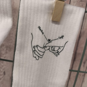 Könnte beinhalten: Weiße Socken mit einer schwarzen Linienzeichnung von zwei Händen, die sich mit einem Herzen in der Mitte halten. Der Text "Markus" und "Tanya" steht über den Händen.