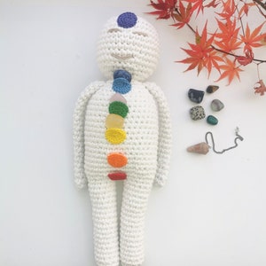 May include: White crocheted doll with rainbow colored stones representing the seven chakras. The doll is lying on a white surface with a small crystal pendulum and other crystals nearby.