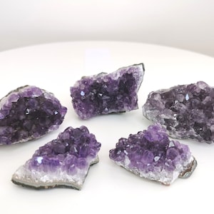 May include: Five amethyst crystal clusters with a deep purple hue. The crystals are clustered together, showcasing their natural formations. The clusters are on a white surface, highlighting the color and texture of the gemstones.