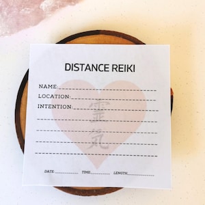 May include: A white card with the text "DISTANCE REIKI" printed in black at the top. The card has lines for writing the name, location, intention, date, time, and length of a Reiki session. A pink heart with a Japanese symbol is in the center of the card.