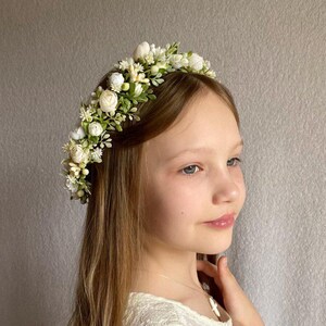 May include: A floral headband featuring an arrangement of small white roses, cream-colored buds, and green foliage. The accessory is worn across the top of the head, showcasing a dense cluster of artificial flowers and leaves set on a thin band.