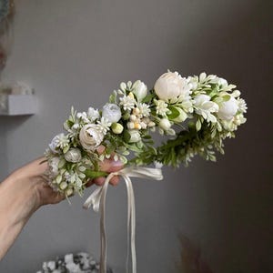 May include: A floral crown featuring an arrangement of white peonies, small white blossoms, and green foliage. The circular headpiece is accented with a thin, light-colored ribbon hanging from the base, creating a delicate and botanical accessory.