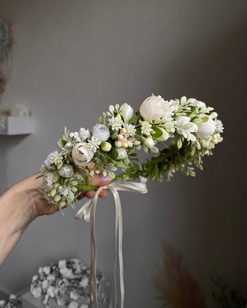 May include: A floral crown featuring an arrangement of white peonies, small white blossoms, and green foliage. The circular headpiece is accented with a thin, light-colored ribbon hanging from the base, creating a delicate and botanical accessory.