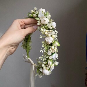 May include: A floral crown featuring white artificial roses, small cream-colored berries, and green foliage arranged on a flexible base. The accessory includes a thin, light-colored ribbon tie at the bottom to secure the circular headpiece in place.