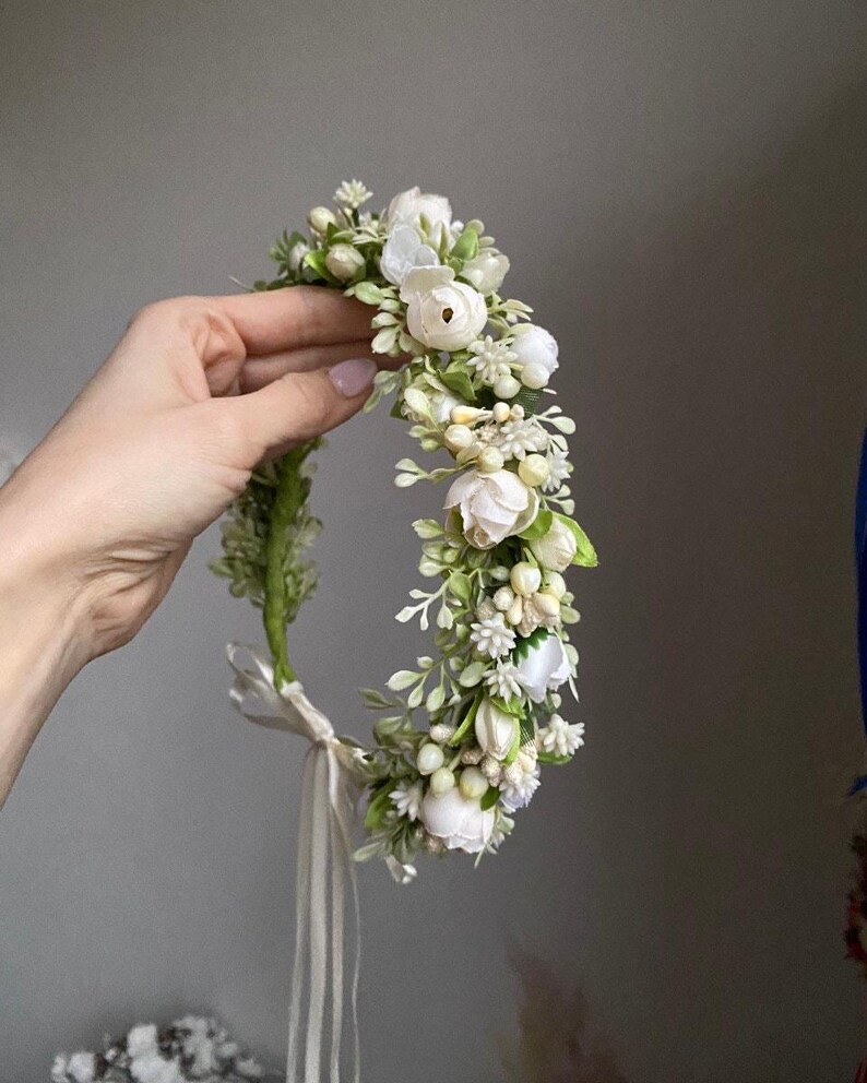 May include: A floral crown featuring white artificial roses, small cream-colored berries, and green foliage arranged on a flexible base. The accessory includes a thin, light-colored ribbon tie at the bottom to secure the circular headpiece in place.