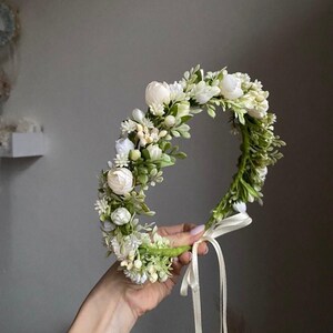 May include: A floral crown featuring white rosebuds, small white blossoms, and green foliage arranged on a circular base. The accessory includes a thin, light-colored ribbon tie hanging from the bottom, providing a secure fit for the wearer.
