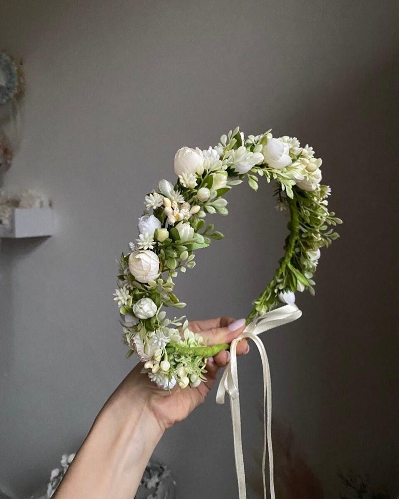 May include: A floral crown featuring white rosebuds, small white blossoms, and green foliage arranged on a circular base. The accessory includes a thin, light-colored ribbon tie hanging from the bottom, providing a secure fit for the wearer.