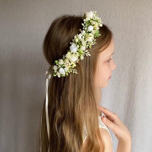 May include: A floral crown featuring an arrangement of small white and cream-colored fabric flowers, buds, and green leaves. The accessory is secured with a long, thin, light-colored ribbon that hangs down from the back of the headpiece.