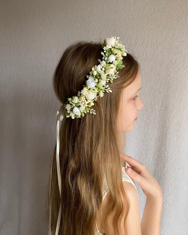 May include: A floral crown featuring an arrangement of small white and cream-colored fabric flowers, buds, and green leaves. The accessory is secured with a long, thin, light-colored ribbon that hangs down from the back of the headpiece.
