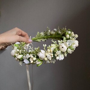 May include: A floral crown featuring white ranunculus flowers, small white blossoms, and green foliage arranged on a circular base. The accessory includes long, thin, light-colored ribbons hanging from the back for adjustment and secure fastening.