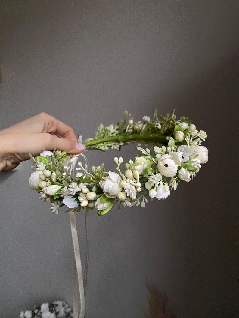 May include: A floral crown featuring white ranunculus flowers, small white blossoms, and green foliage arranged on a circular base. The accessory includes long, thin, light-colored ribbons hanging from the back for adjustment and secure fastening.