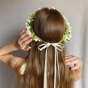 May include: A floral crown featuring small white and yellow blossoms with green foliage, secured at the back with a long, thin cream-colored ribbon tied in a bow. The accessory is positioned on long brown hair, with the ribbon tails hanging down.