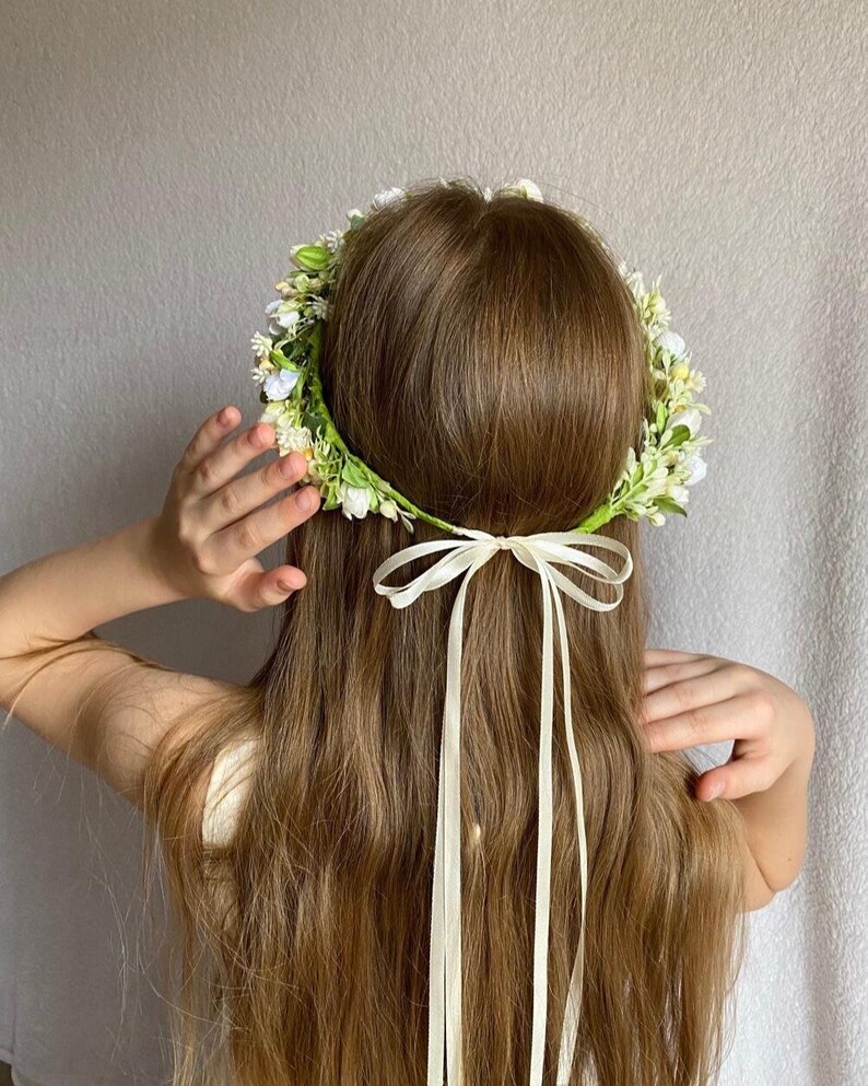 May include: A floral crown featuring small white and yellow blossoms with green foliage, secured at the back with a long, thin cream-colored ribbon tied in a bow. The accessory is positioned on long brown hair, with the ribbon tails hanging down.