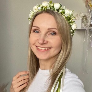 May include: A floral crown featuring small white blossoms and green foliage, accented with delicate white buds. The headpiece includes long, trailing ribbons in shades of cream and olive green that hang down from the sides of the circular arrangement.