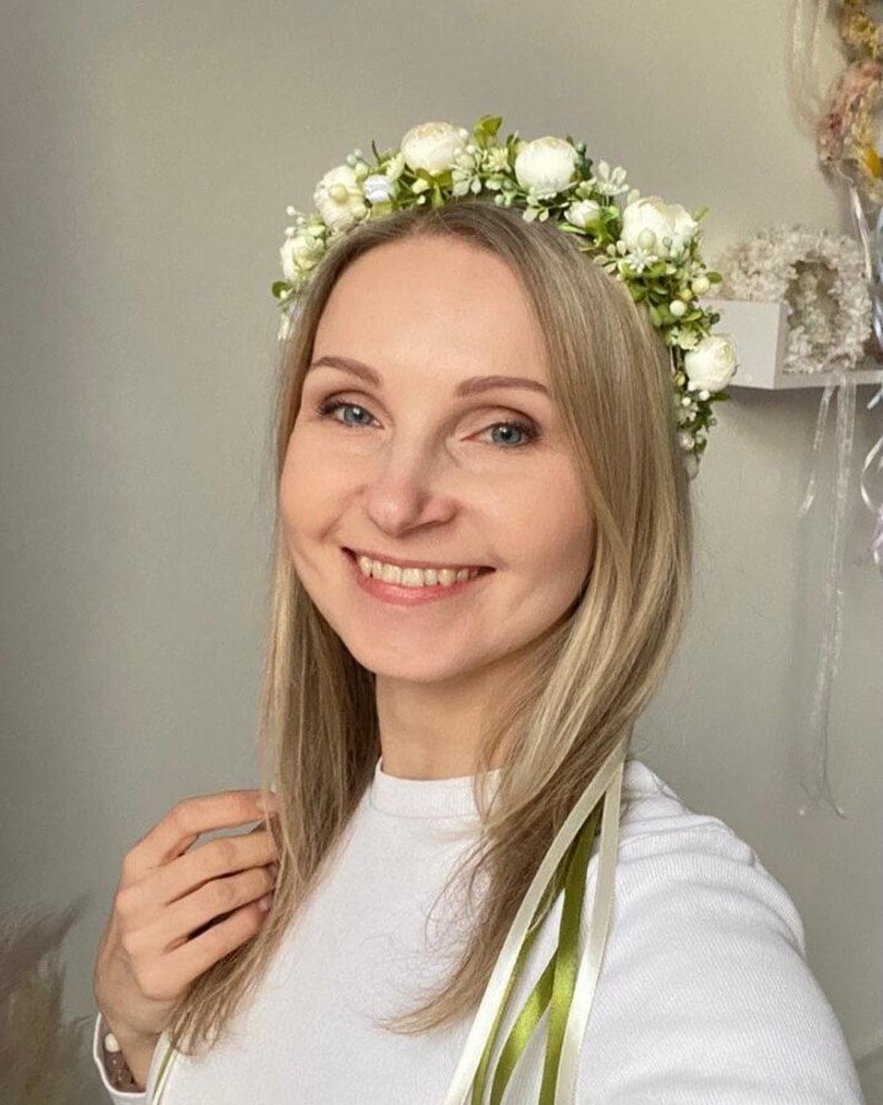 May include: A floral crown featuring small white blossoms and green foliage, accented with delicate white buds. The headpiece includes long, trailing ribbons in shades of cream and olive green that hang down from the sides of the circular arrangement.
