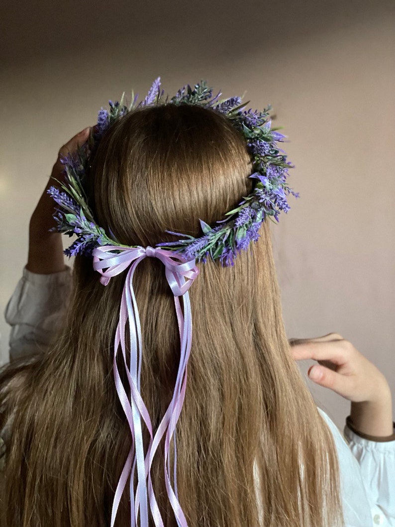 May include: A purple flower crown with a lavender ribbon tied in a bow. The crown is made of artificial flowers and is worn on a person's head.