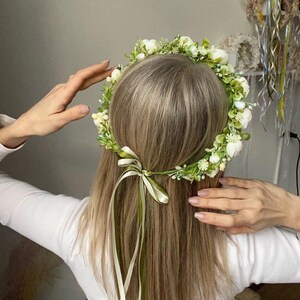 May include: A floral crown featuring small white blossoms and green foliage arranged in a circular band. The accessory is secured at the back with two thin ribbons, one in a light cream shade and the other in a muted olive green, tied in a bow.