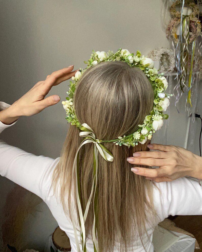 May include: A floral crown featuring small white blossoms and green foliage arranged in a circular band. The accessory is secured at the back with two thin ribbons, one in a light cream shade and the other in a muted olive green, tied in a bow.
