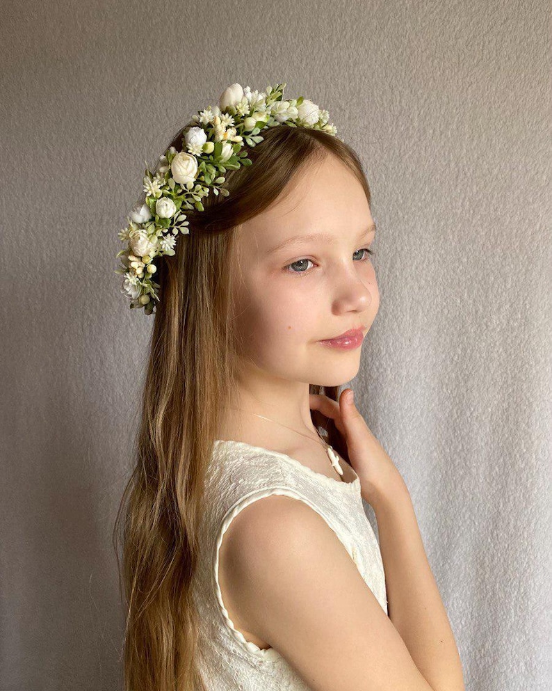 May include: A floral headband featuring an arrangement of small white roses, cream-colored buds, and green foliage. The accessory is worn across the top of the head, showcasing a dense cluster of artificial flowers and leaves set on a thin band.