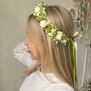 May include: A floral crown featuring white roses, small white buds, and green foliage. The accessory is secured with long, flowing olive green and white satin ribbons that hang down from the back, creating a natural and botanical appearance.