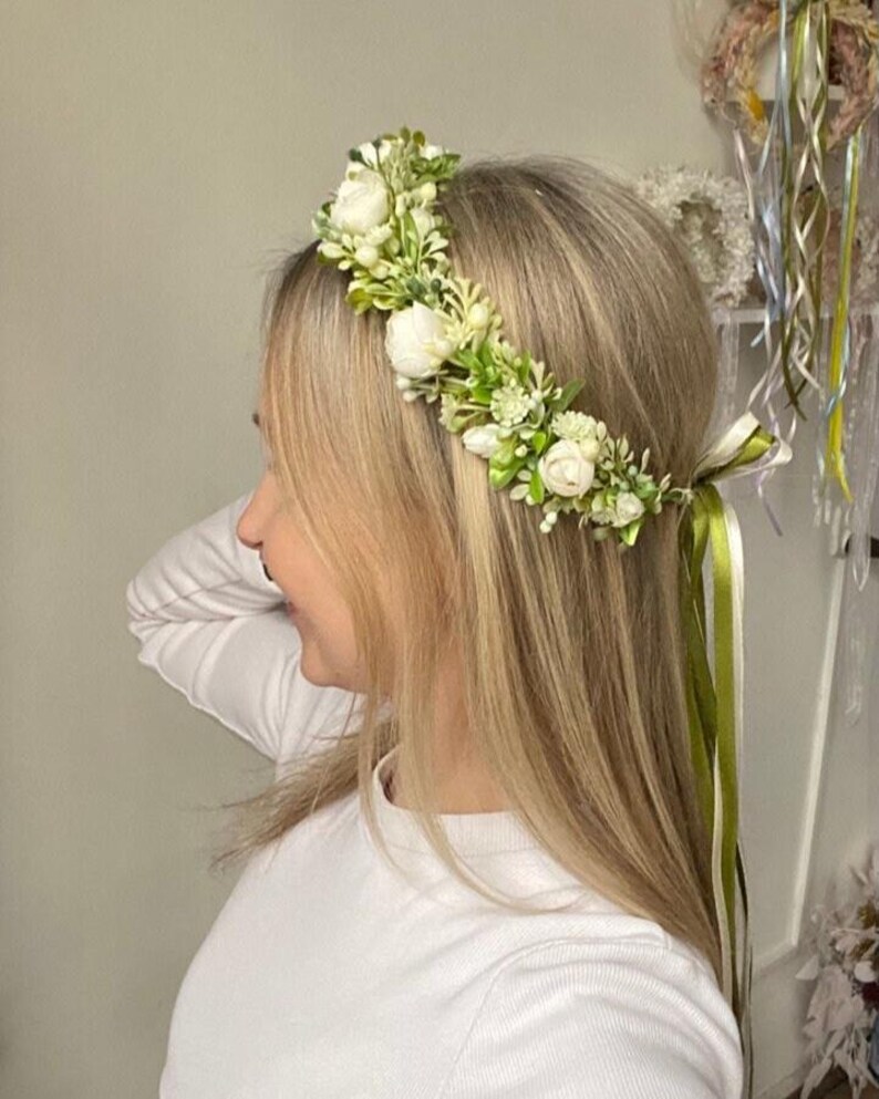 May include: A floral crown featuring white roses, small white buds, and green foliage. The accessory is secured with long, flowing olive green and white satin ribbons that hang down from the back, creating a natural and botanical appearance.