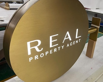 May include: A round, gold-colored sign with the word "REAL" and the text "PROPERTY AGENT" in white lettering.