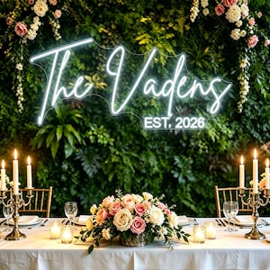 May include: A wedding reception table setting with a floral centerpiece, candles, and a neon sign that reads "The Vadens EST. 2026" against a backdrop of greenery and flowers. The table is set for a celebration.