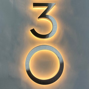 Könnte beinhalten: Hausnummer 30 aus gebürstetem Metall mit warmer LED-Hintergrundbeleuchtung. Die Zahlen sind an einer hellgrauen Wand angebracht. Das Design zeichnet sich durch eine moderne, minimalistische Ästhetik aus, geeignet für den Außen- oder Innenbereich.