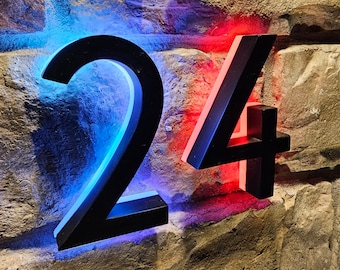 May include: Black metal house numbers 24 with blue and red LED lights illuminating the numbers against a stone wall background.