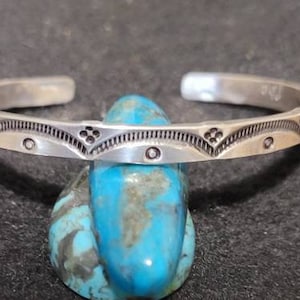 May include: A silver cuff bracelet with an engraved geometric pattern. The bracelet is displayed on a blue and turquoise stone against a dark background. The bracelet's design includes small circles and floral-like shapes.