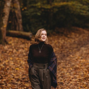 May include: A woman wearing a brown turtleneck sweater, a plaid scarf, and brown tweed pants. She is standing in a forest with fallen leaves on the ground.