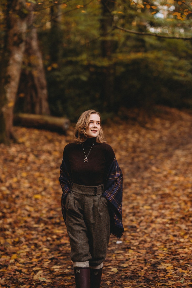 May include: A woman wearing a brown turtleneck sweater, a plaid scarf, and brown tweed pants. She is standing in a forest with fallen leaves on the ground.